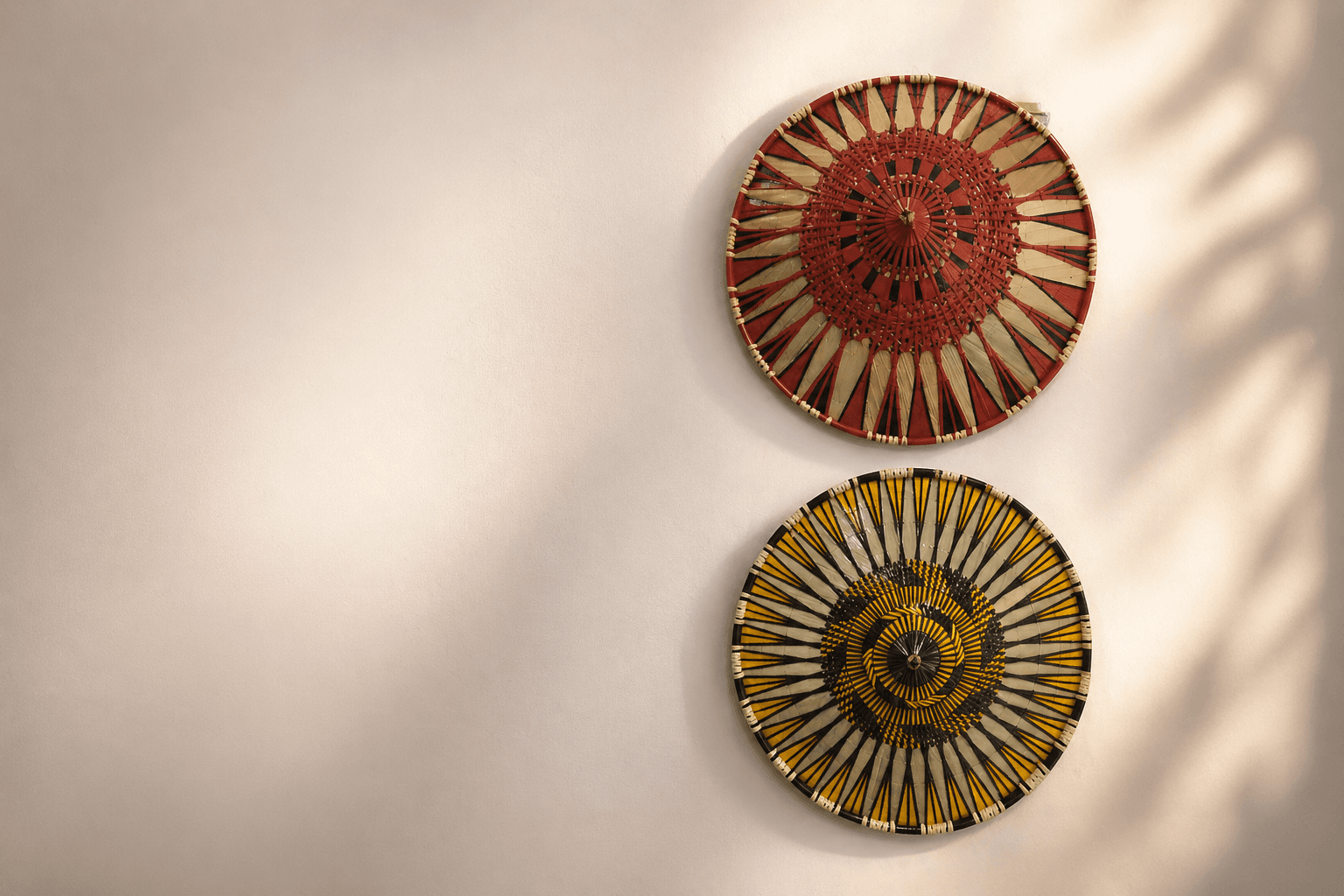 Two intricately patterned woven conical hats mounted on a neutral wall with soft, dappled shadows.